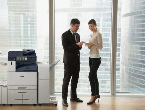 Due persone in un ufficio in abiti da lavoro che discutono di documenti sono in piedi accanto a una stampante multifunzione Xerox® AltaLink® serie B8200